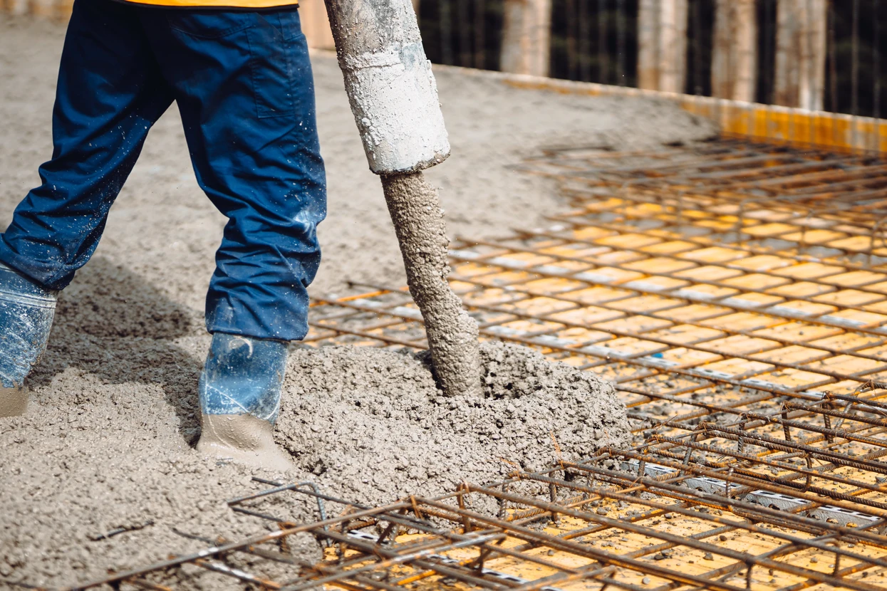 Chantier de construction en extérieur avec ouvrier en combinaison bleue coulant du béton liquide via une goulotte sur ferraillage
